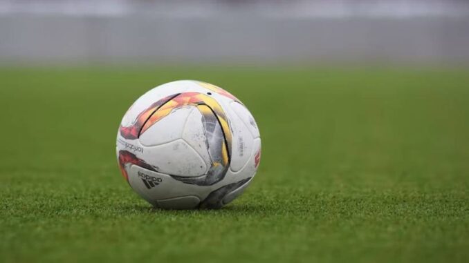 Balón de fútbol blanco con estampado rojo y negro sobre el césped verde de un campo de juego.